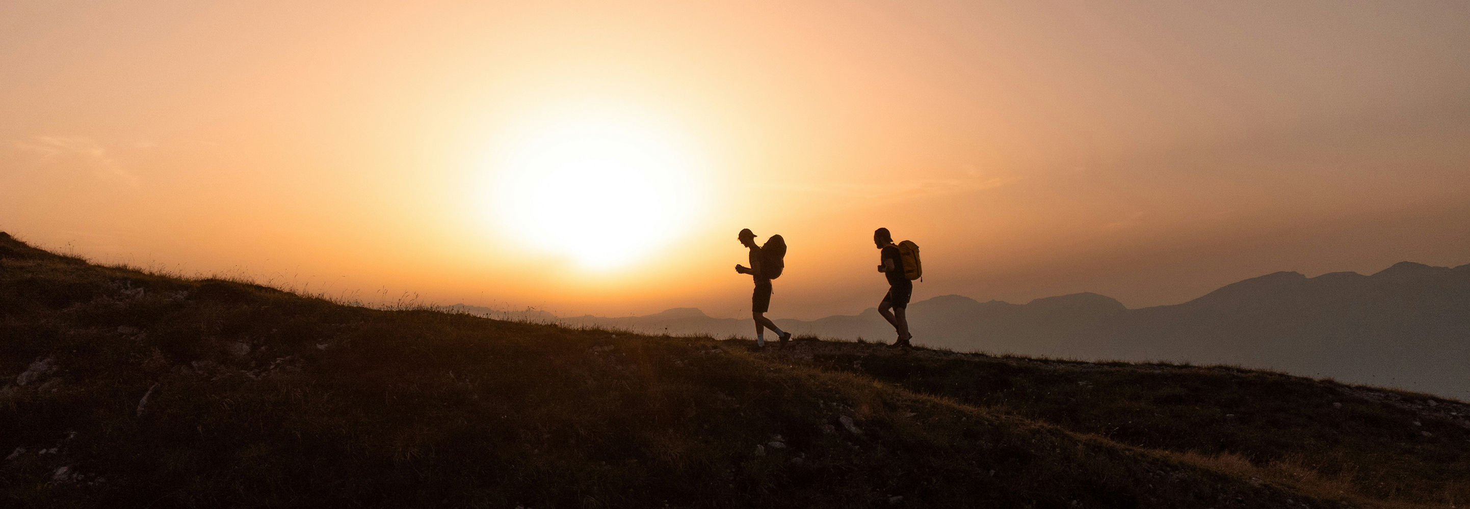 Two hikers with backpacks walking on a hill during sunset with mountains in the background.