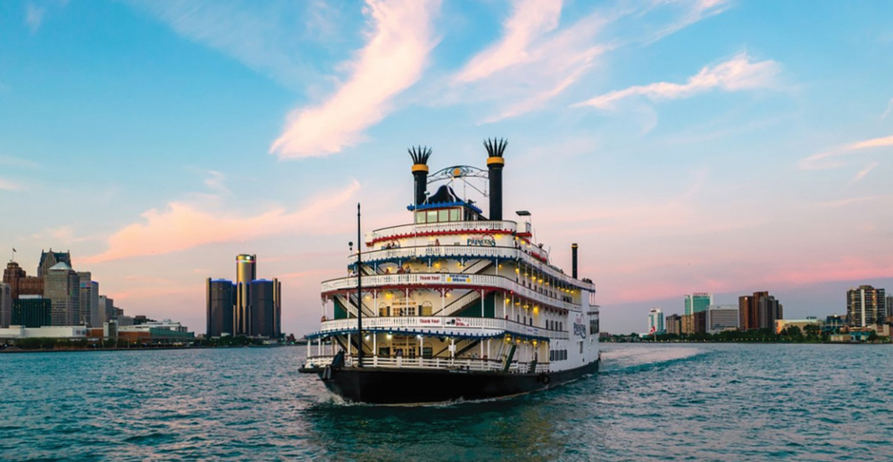 Large white riverboat cruising on a river with a city skyline at sunset in the background.