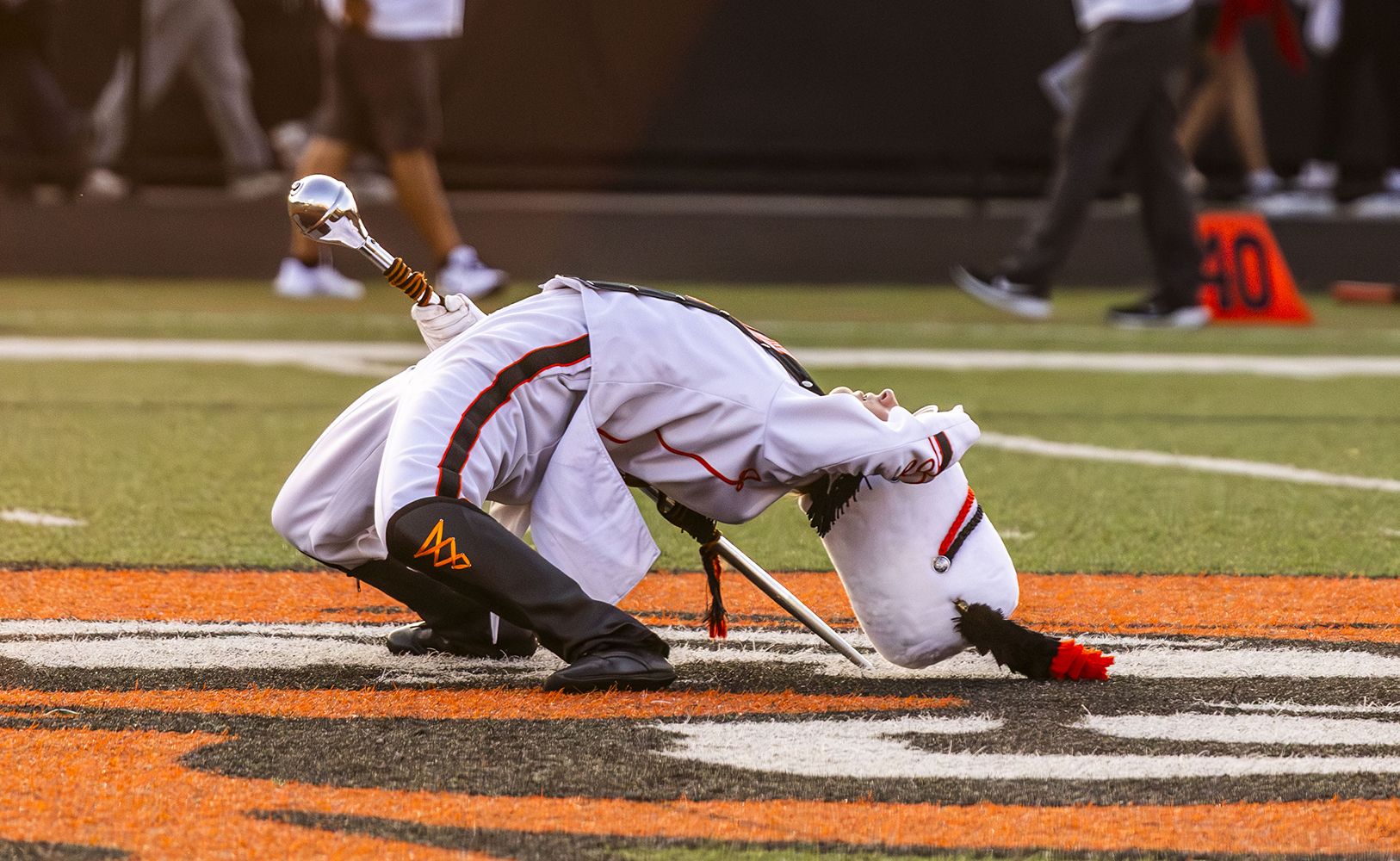 Marching band member in white uniform and tall hat bending backward on a football field holding a silver mace.