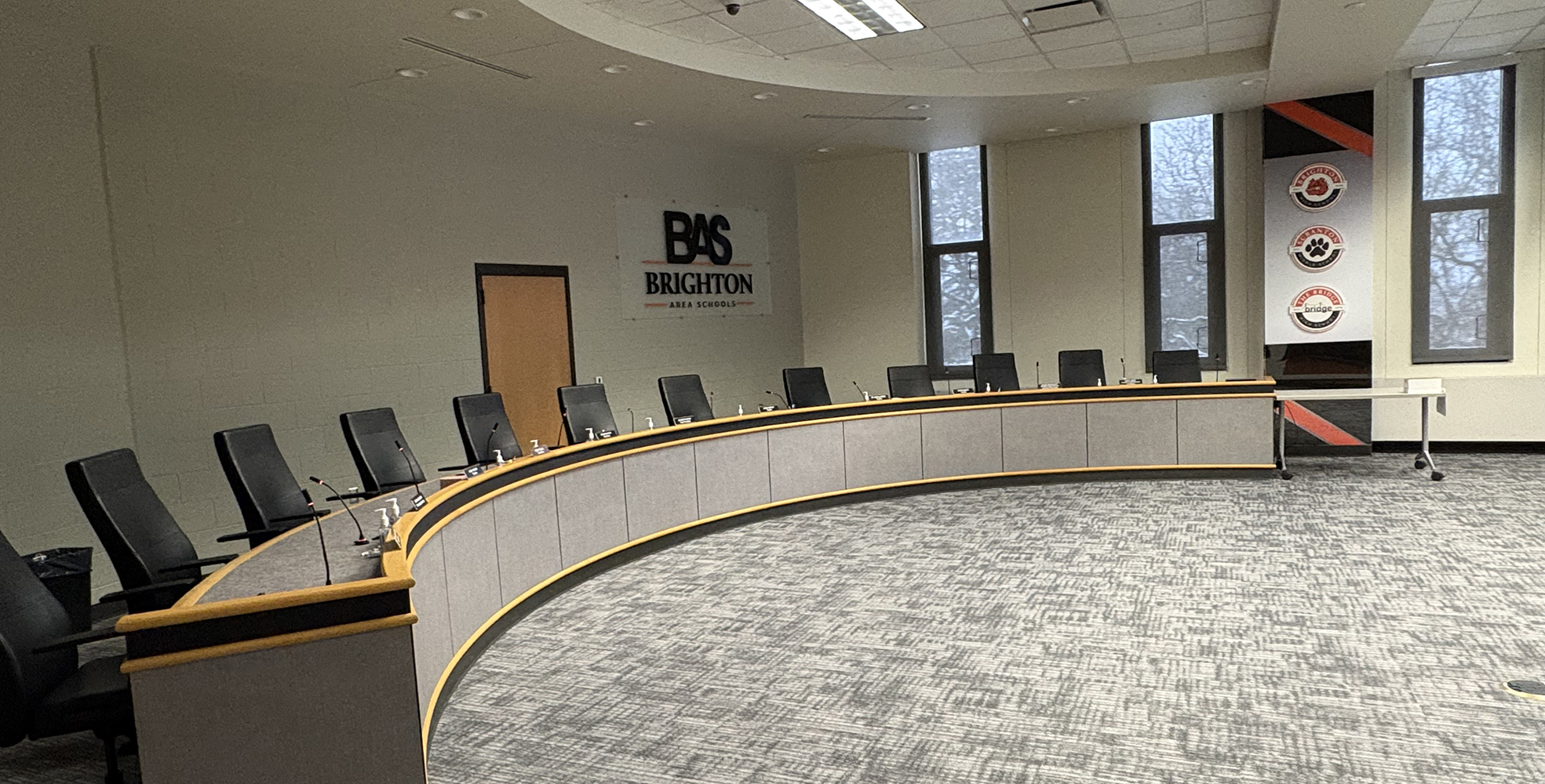 Curved conference table with black chairs and microphones in a boardroom with Brighton Area Schools signage on the wall.