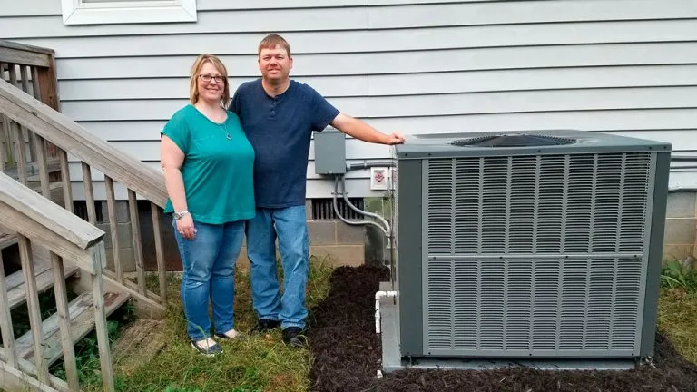 A smiling couple stands outside their house next to a newly installed large HVAC unit.