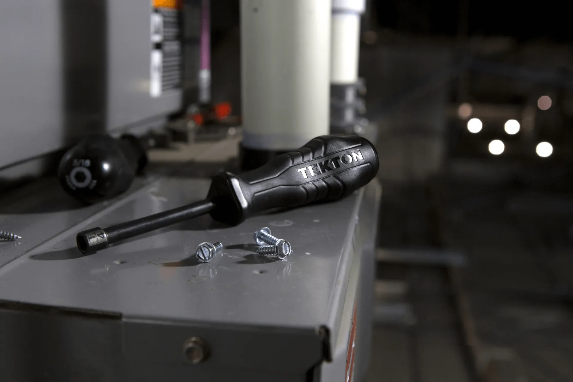 Close-up of a black TEKTON screwdriver resting on a metal surface next to a few loose screws, with blurred industrial equipment in the background.