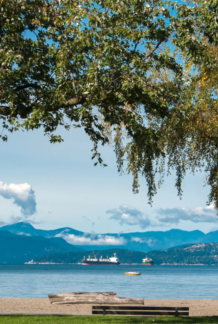 Scenic view of English Bay beach with driftwood, ocean sailboats, and North Shore mountains, highlighting the natural surroundings near Point Grey Mews Vancouver.