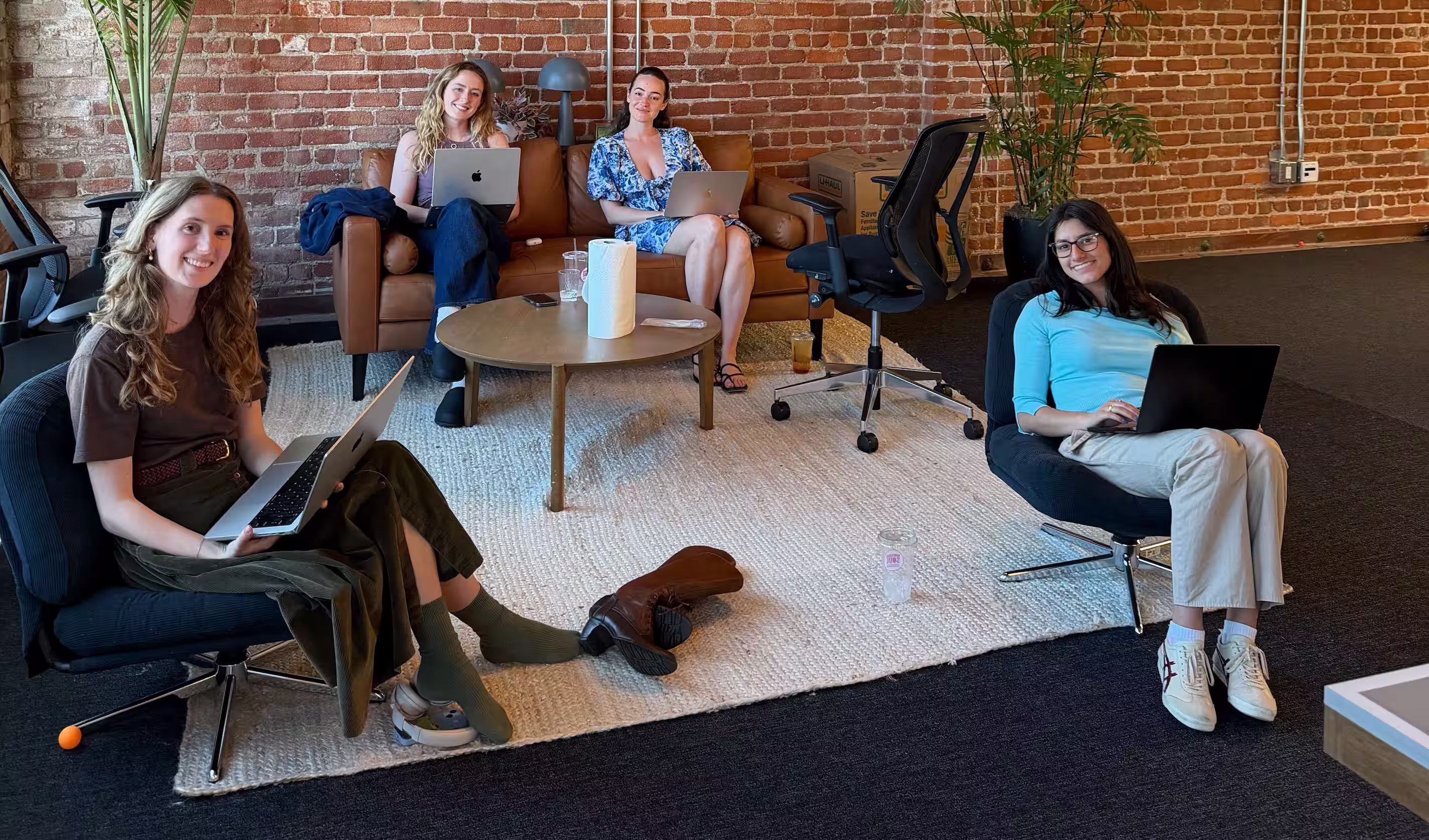 Four team members working on laptops in a relaxed coworking lounge with exposed brick walls, wooden beam ceilings, and warm ambient lighting.