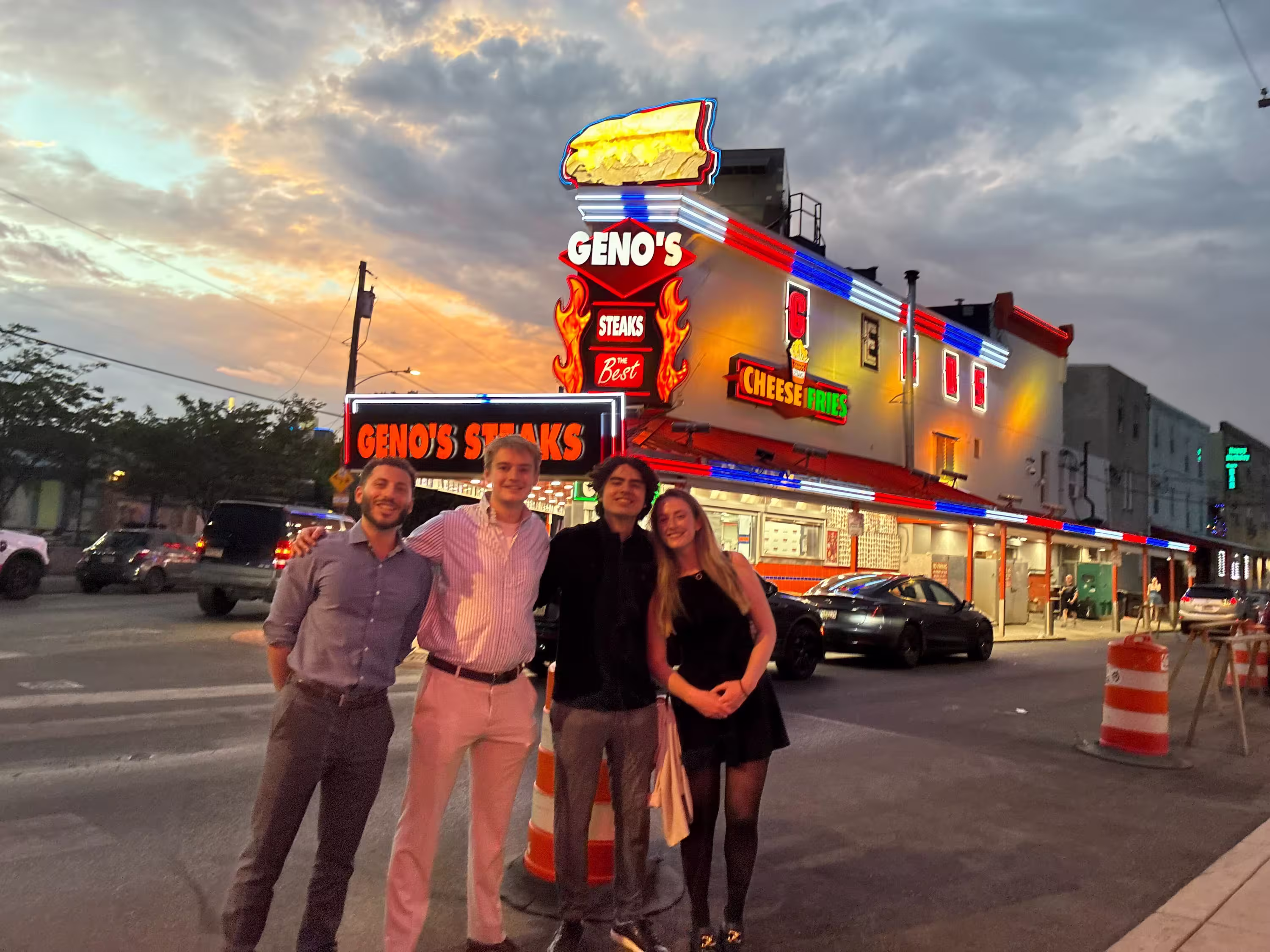 Four team members smiling together in front of Geno's Steaks in Philadelphia at dusk, with the restaurant's iconic neon signage lit up behind them.