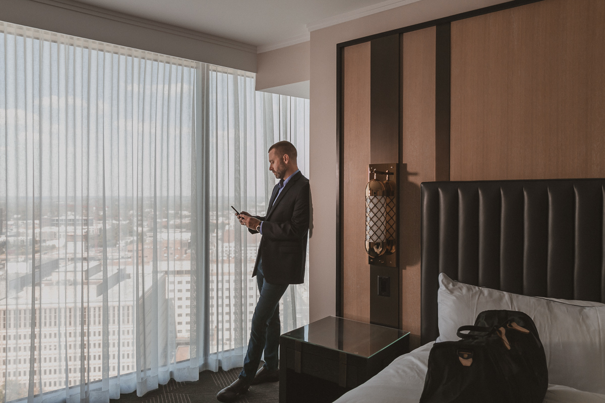 commercial photography of a man looking at his phone in a hotel room with an overnight bag on the bed