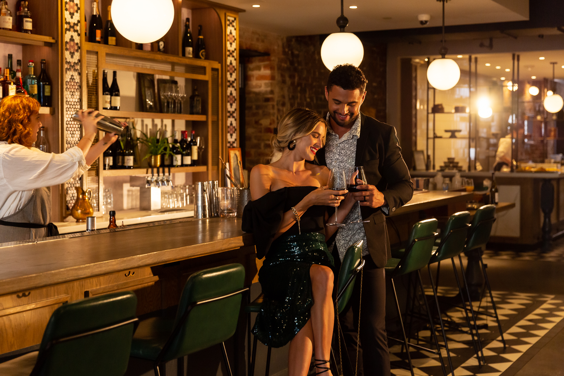 commercial photography of a couple sitting at the bar drinking while a bartender mixes drinks