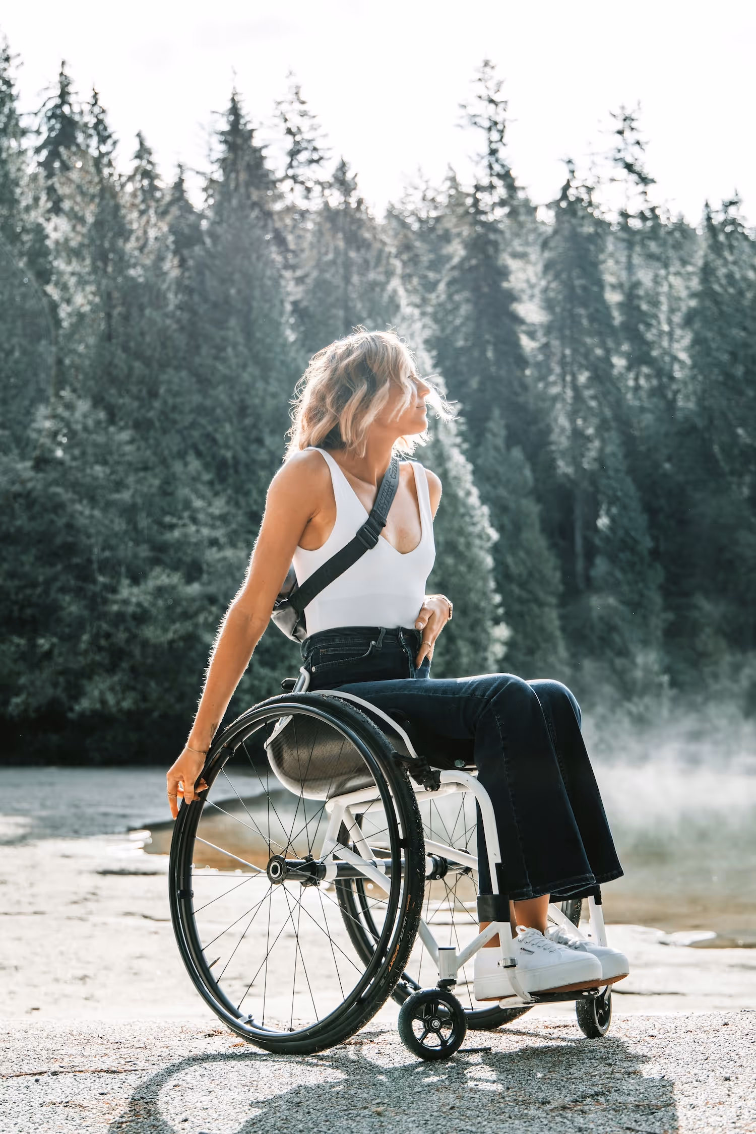 Woman with blonde hair in a wheelchair by a lake, surrounded by forest, wearing a white tank top and dark jeans.
