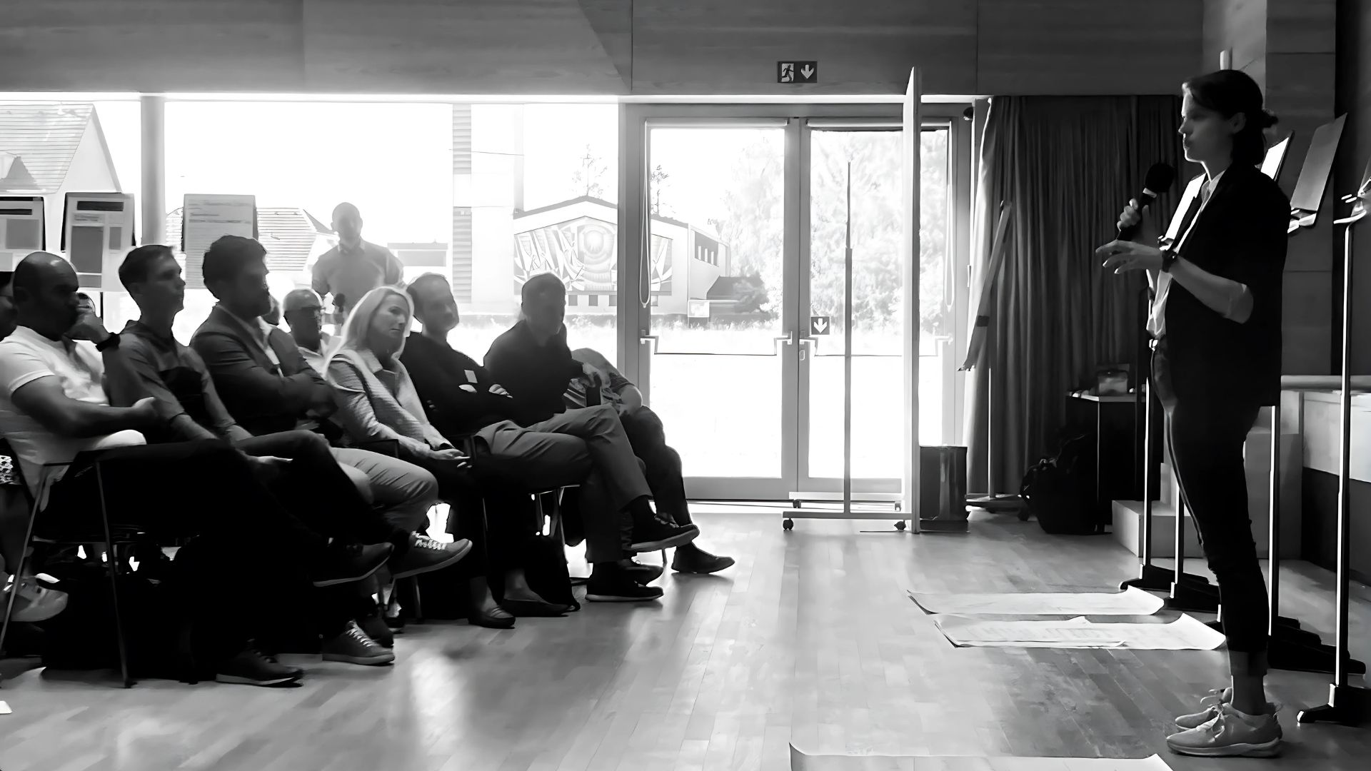 Konzept-Workshop von sisigrant: Iris Borovčnik spricht mit Mikrofon vor einer Gruppe, die im Halbkreis sitzt und zuhört.