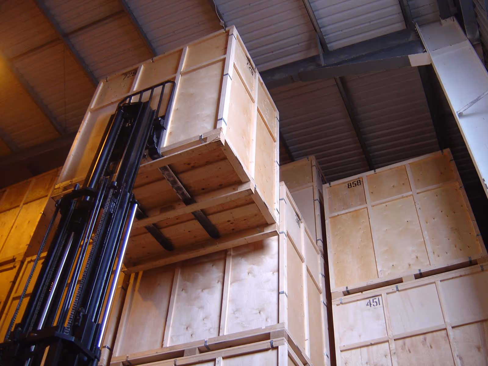 A forklift lifts large wooden crates in a warehouse, stacking them high near the ceiling.