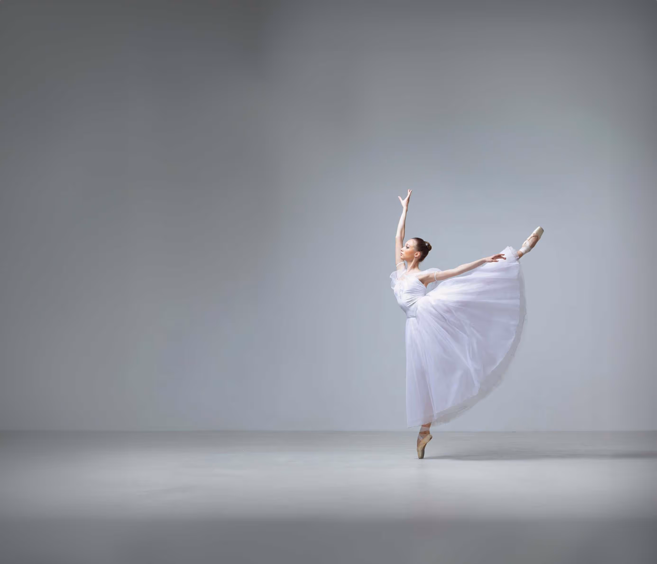 Ballet dancer in white dress performs a graceful arabesque on a bare stage.