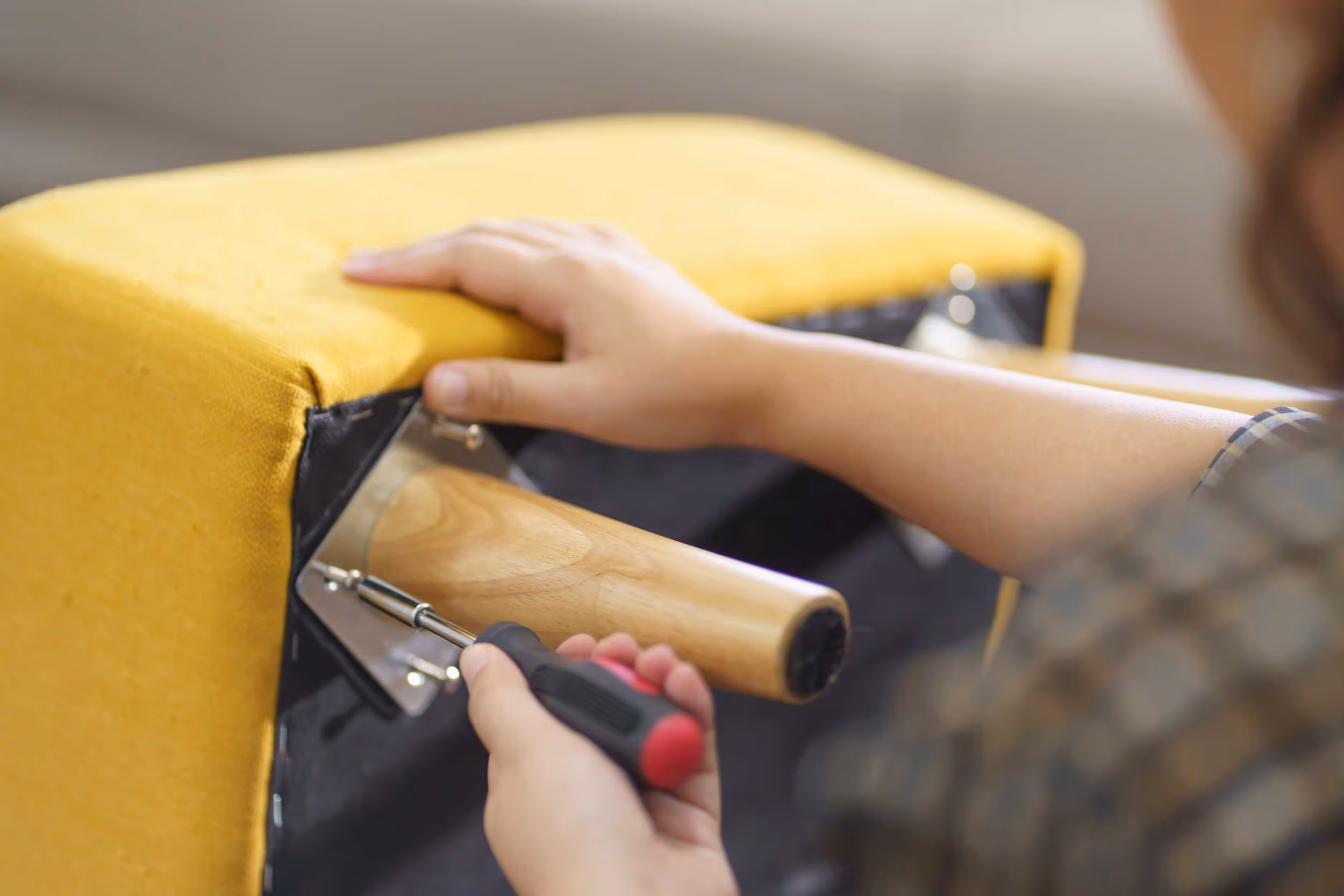 A person tightens a screw on a yellow upholstered chair using a screwdriver.