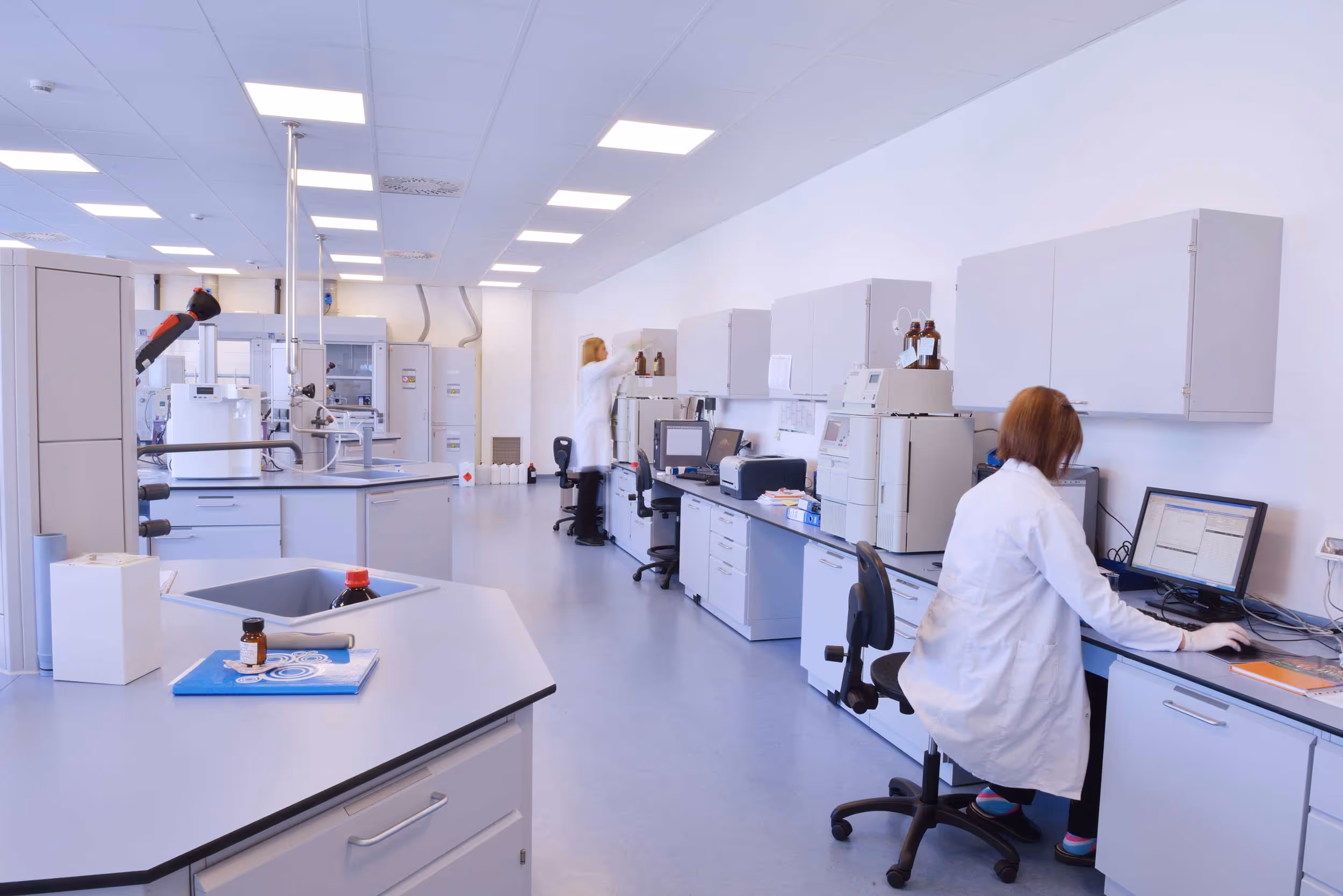 Three scientists in lab coats work at computers and equipment in a bright laboratory.