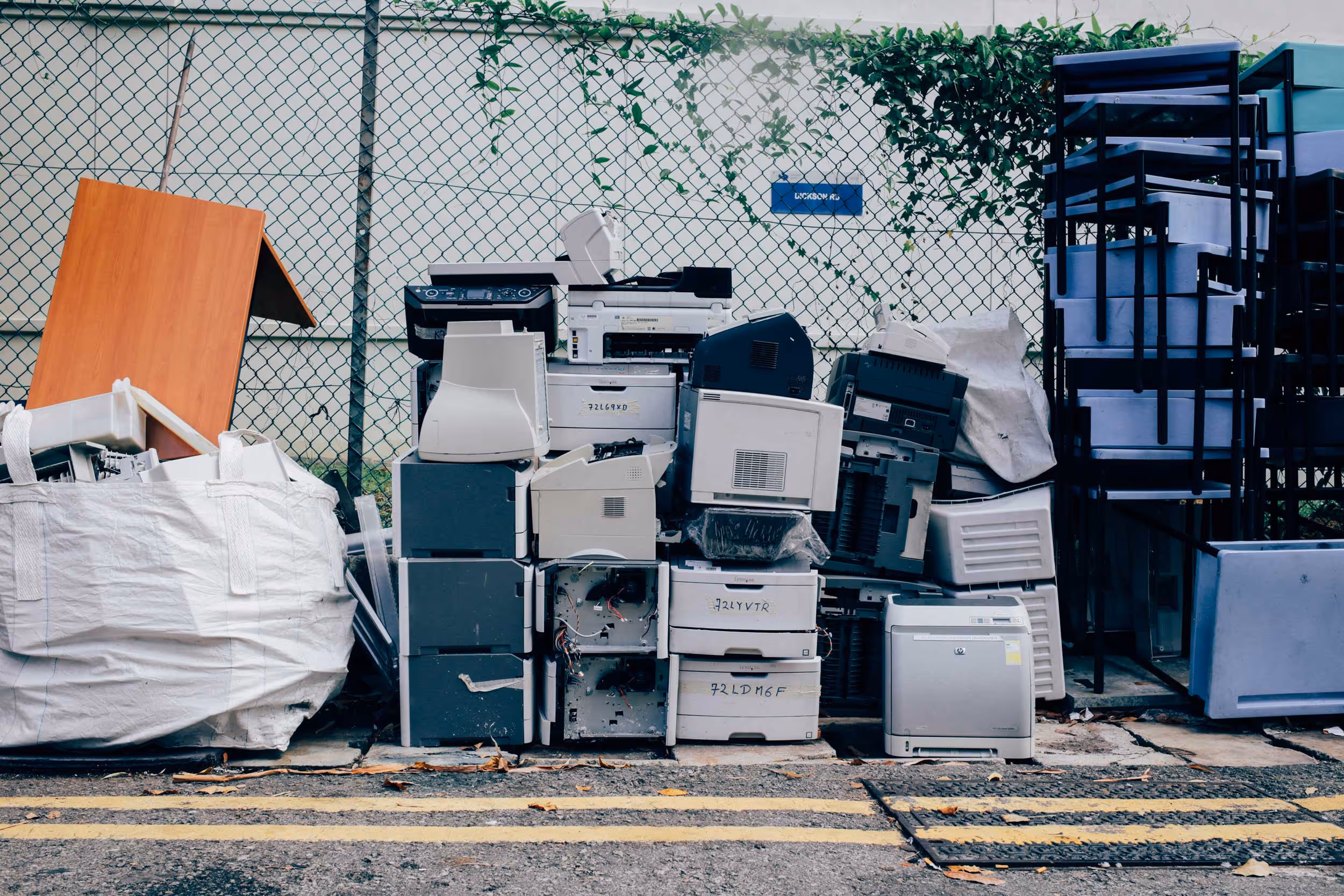A pile of discarded electronic devices, including printers and copiers, stacked against a chain-link fence.