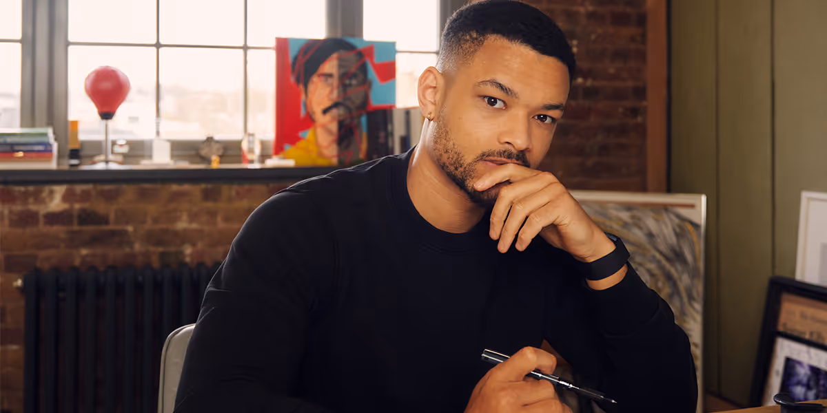 Steven Bartlett wearing a black sweater sits thoughtfully at a desk holding a pen, with framed artwork and a window in the background.