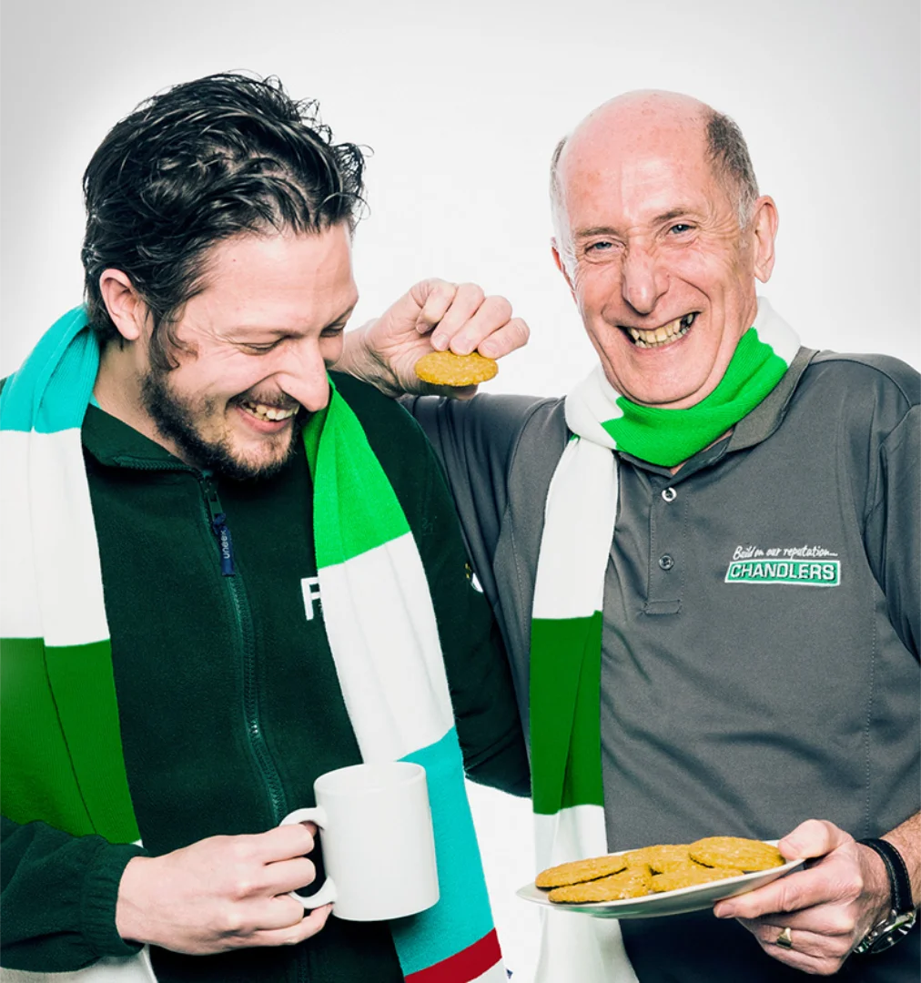 Two happy older men with tea, biscuits and an IBGM branded scarf each laughing together.  