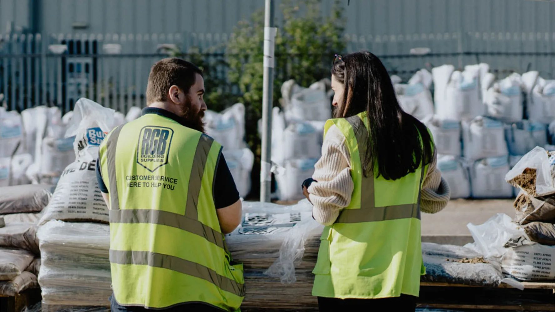 A man and a woman in high visibility vests outside a builders merchant talking while facing away from the camera