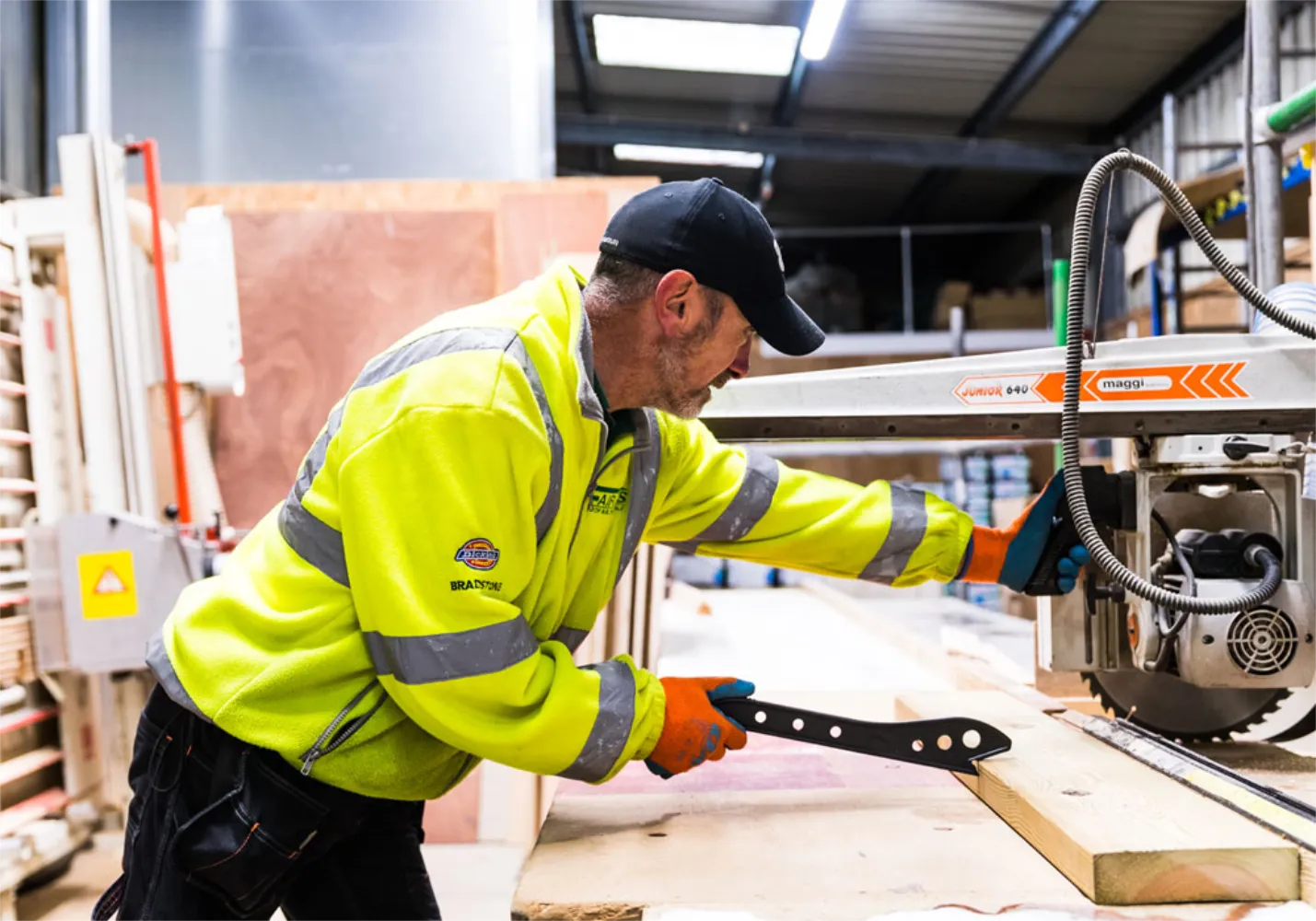 Man in a high visibility jacket with a black cap on pushing some timber through a table saw with a plastic tool.