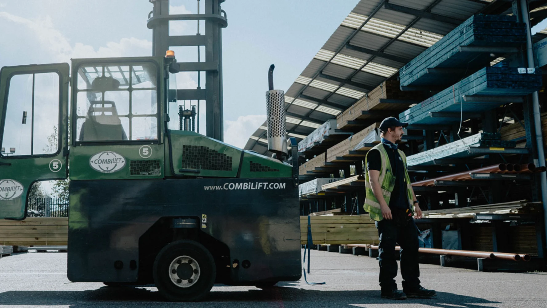 Man outside in a high visibility vest looking away from a large forklift.