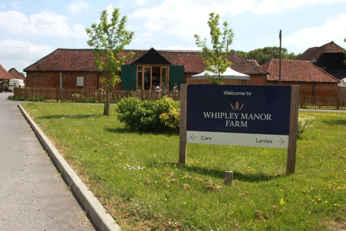 Whipley Manor Farm entrance sign in blue and cream outside of a building next to a road.
