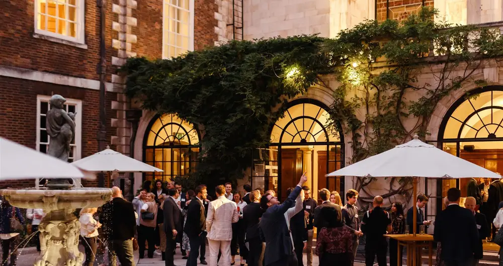 Evening outdoor gathering with people socializing near a courtyard fountain and arched windows with warm lighting.