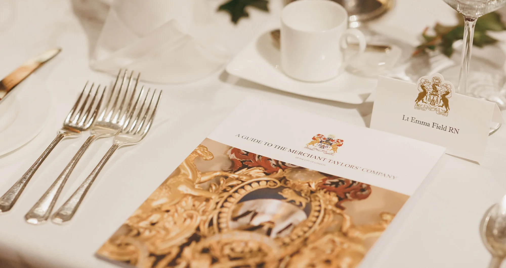 Formal table setting with three forks, a knife, a white cup on a saucer, and a brochure titled 'A Guide to the Merchant Taylors' Company' alongside a name card for Lt Emma Field RN.