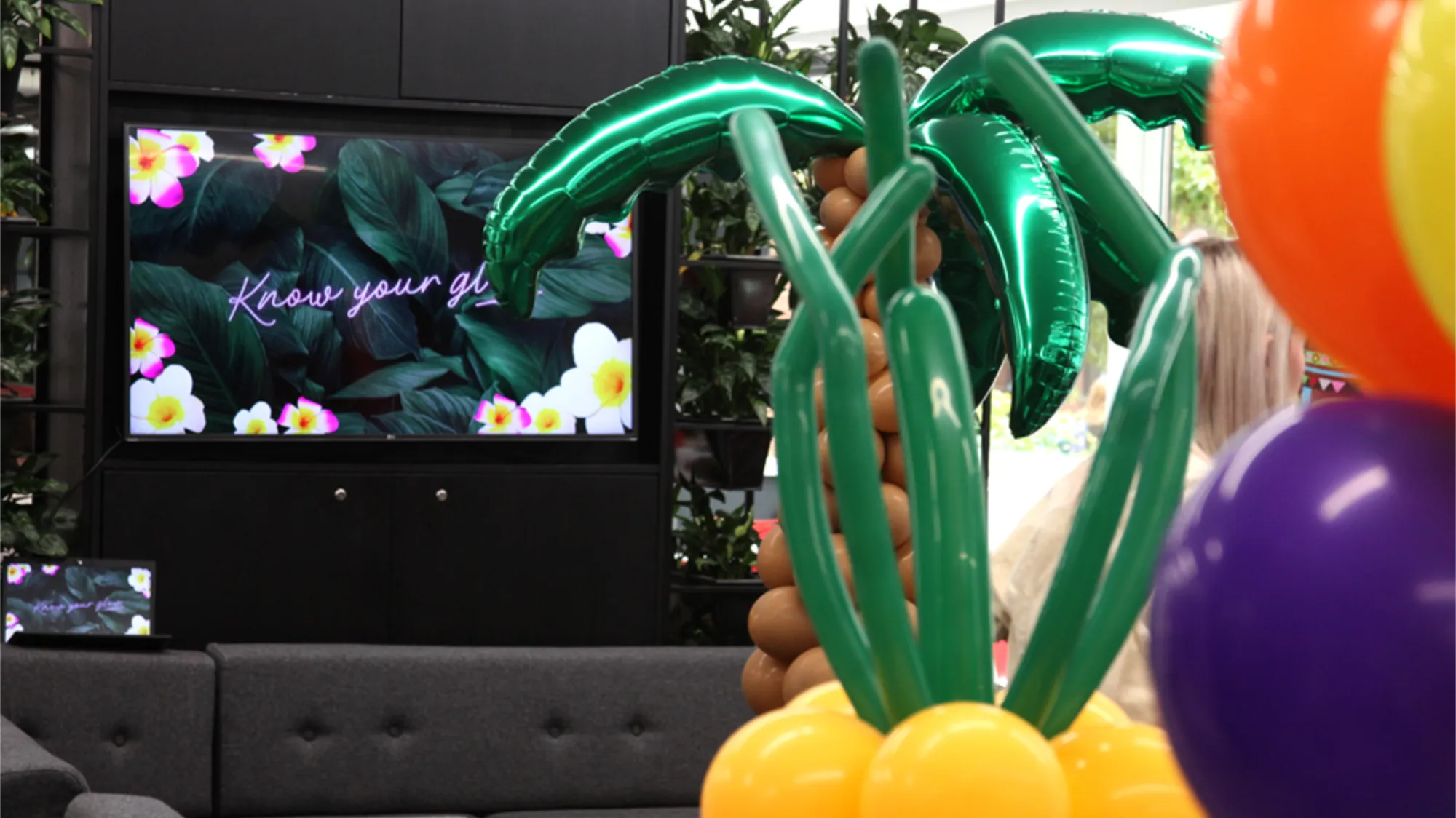 Indoor setting with balloon decorations shaped like a palm tree and colorful balloons, a TV screen displaying floral design with text, and a grey sofa.