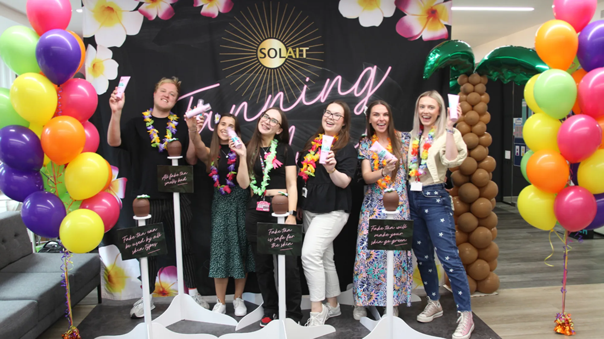 Six people wearing flower leis holding skincare tubes, standing in front of a festive backdrop with bright balloons and inflatable palm tree at a Solait tanning event.
