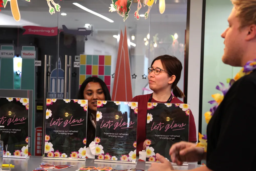 Two women behind a counter with 'let's glow' promotional signs and a man with a flower lei in the foreground.