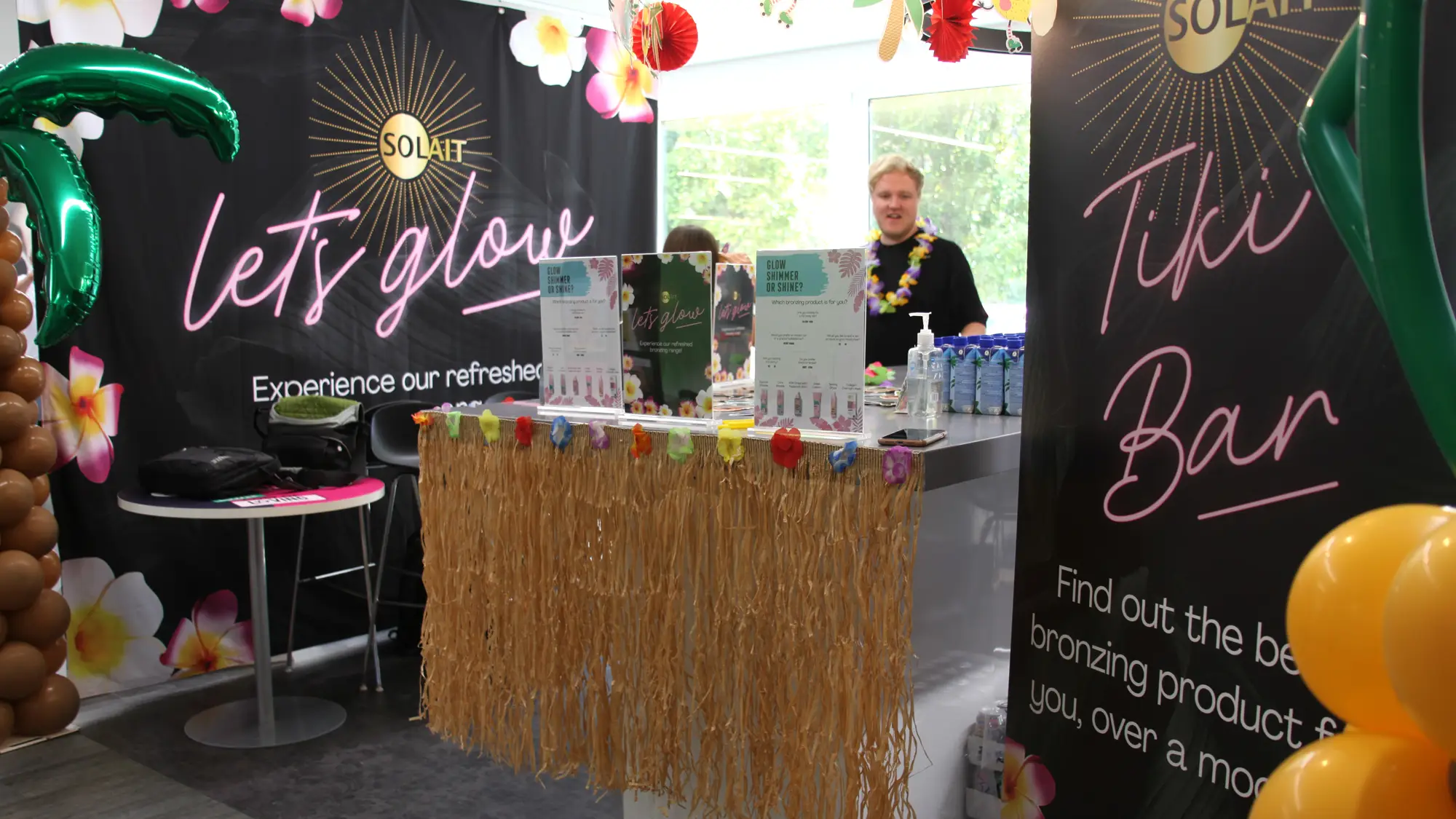 Indoor tiki bar decorated with tropical balloons, floral garlands, and signs promoting 'Let's Glow' and bronzing products with people behind the counter.
