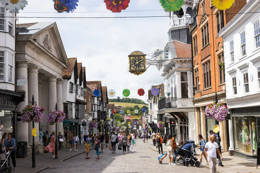 Busy pedestrian street in a historic town with hanging flower baskets, colorful flower decorations overhead, and a prominent clock on an ornamental bracket.