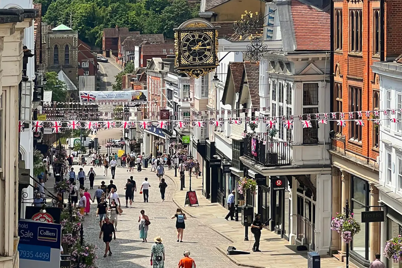 Busy cobblestone street in a historic town with hanging Union Jack flags and a large ornate clock overhead.