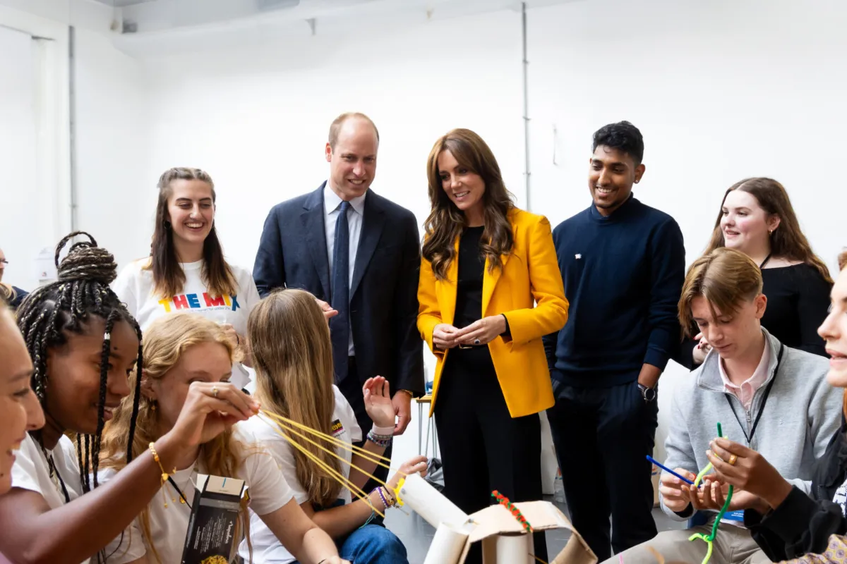 A group of young people interacting with Prince William and Princess Kate, one wearing a yellow blazer, in a bright room while working on a project involving rubber bands and small structures.