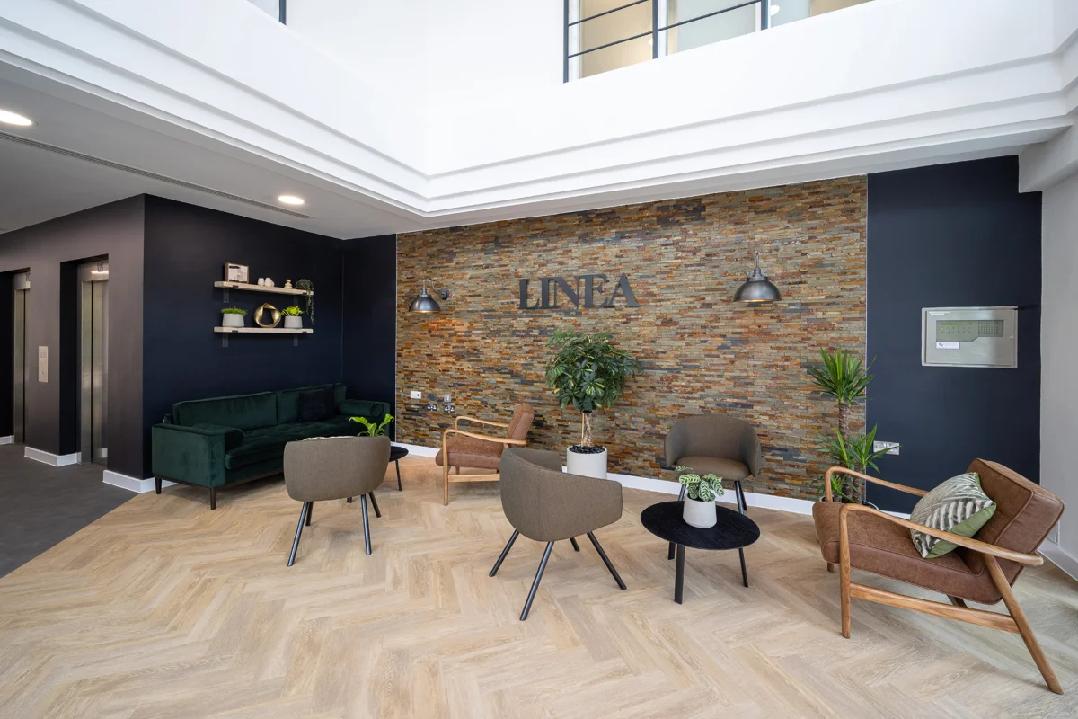 Modern office lounge area with a stone accent wall reading 'LINEA,' surrounded by various chairs, a green velvet sofa, and plants under natural light from a skylight.