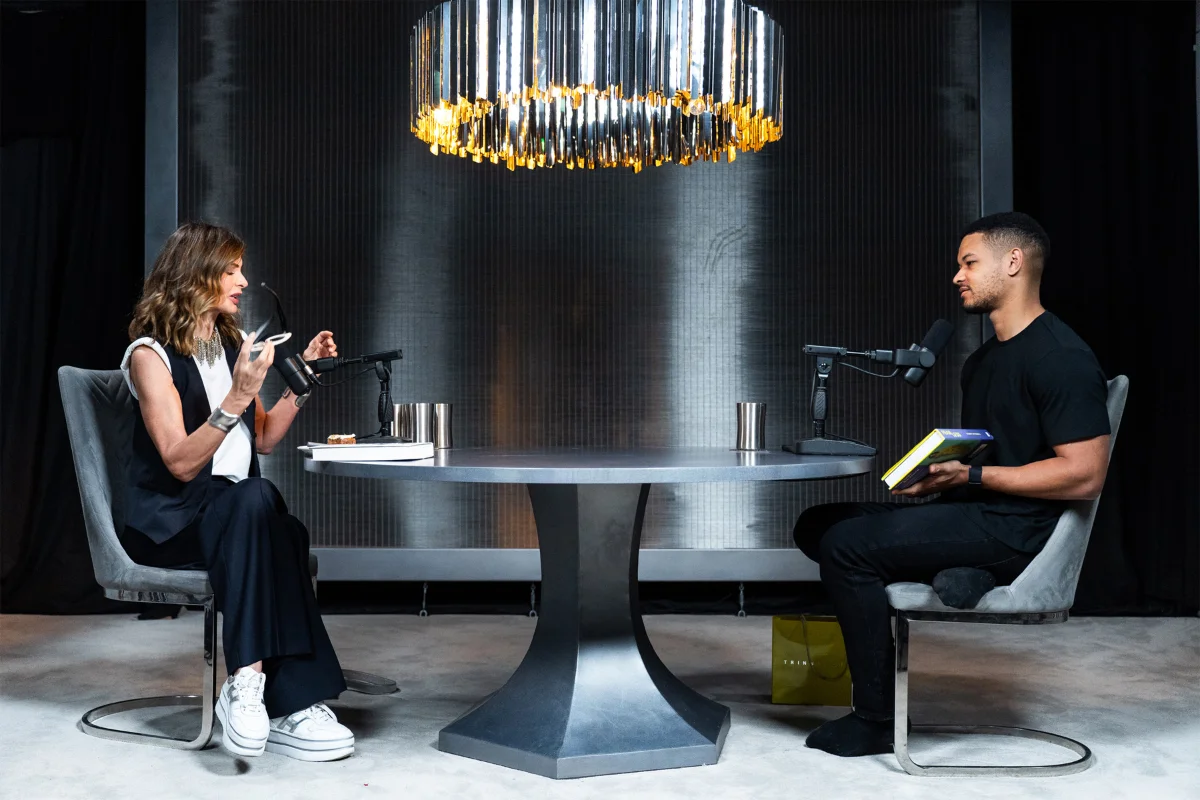 Steven Bartlett and a woman seated across from each other at a round table with microphones, recording a podcast