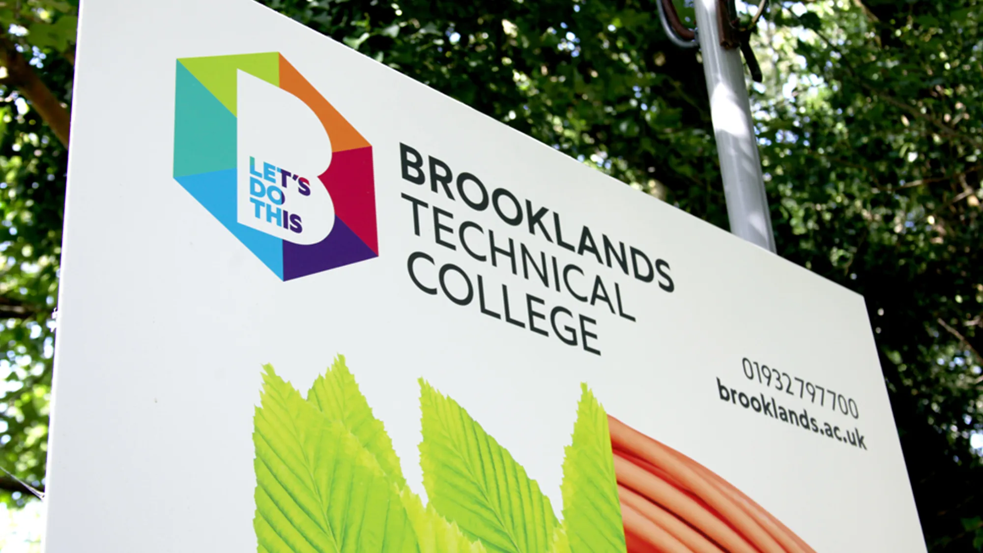 Signboard for Brooklands Technical College with colorful logo and contact information on a background of green trees.