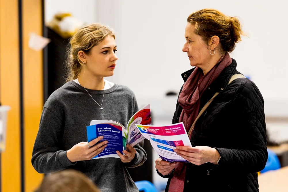 Mother and daughter facing each other and holding colourful Brooklands College prospectus', engaged in conversation indoors.