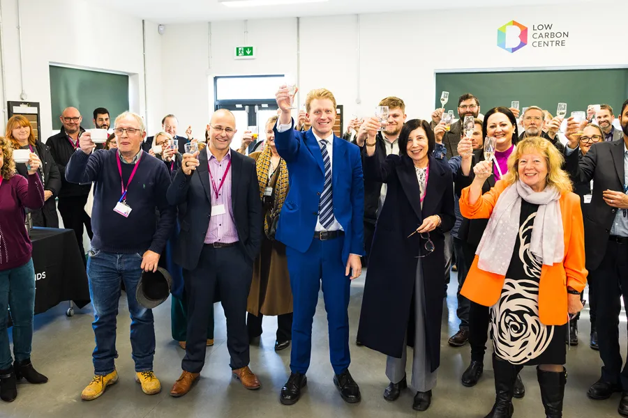 Group of smiling people raising glasses in a toast at an indoor event with Low Carbon Centre logo on the wall.