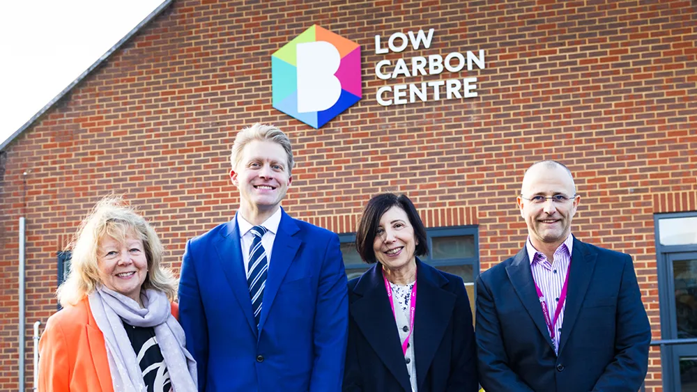 Four people standing and smiling in front of a brick building with a colourful Brooklands College sign reading Low Carbon Centre.