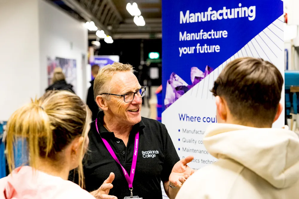 Man wearing glasses and a Brooklands College shirt explains manufacturing career options to two young people near a manufacturing information board.