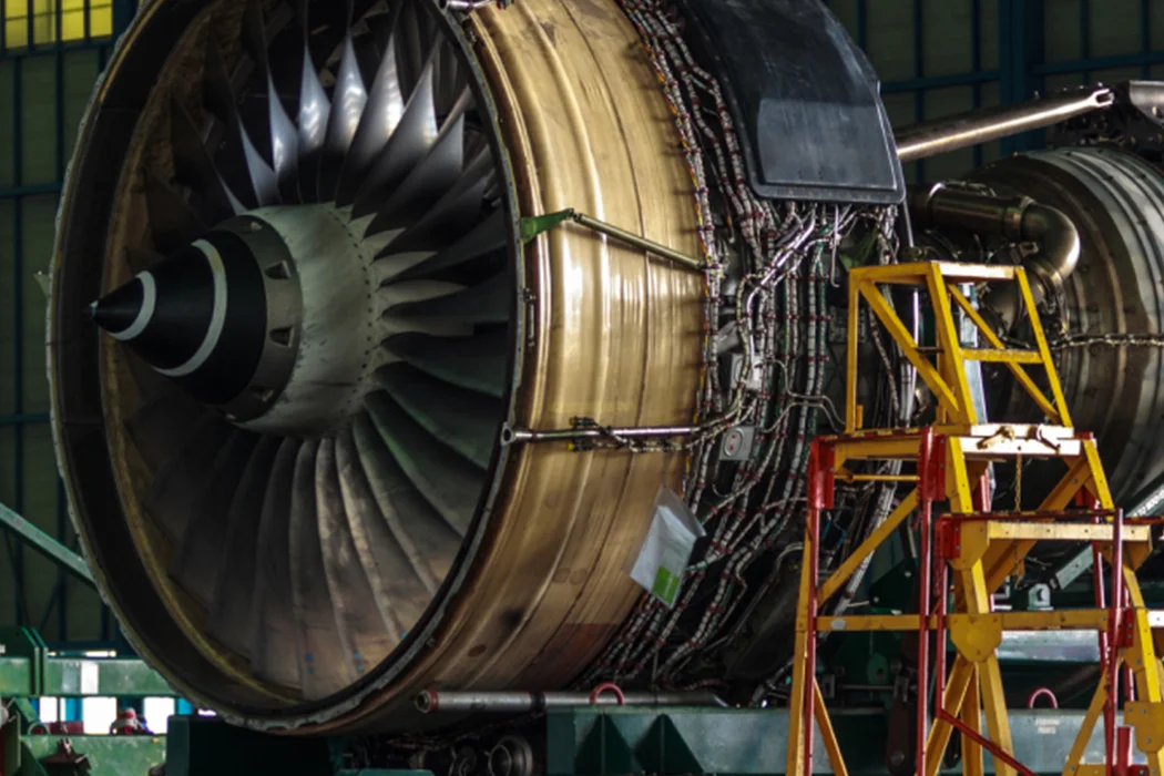 Close-up of a large aircraft jet engine with visible turbine blades and wiring, situated in a maintenance facility with a yellow ladder beside it.