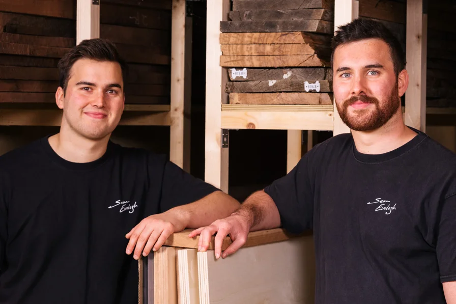 Two men in black Sean Evely t-shirts standing in a woodworking shop with stacked wood behind them.