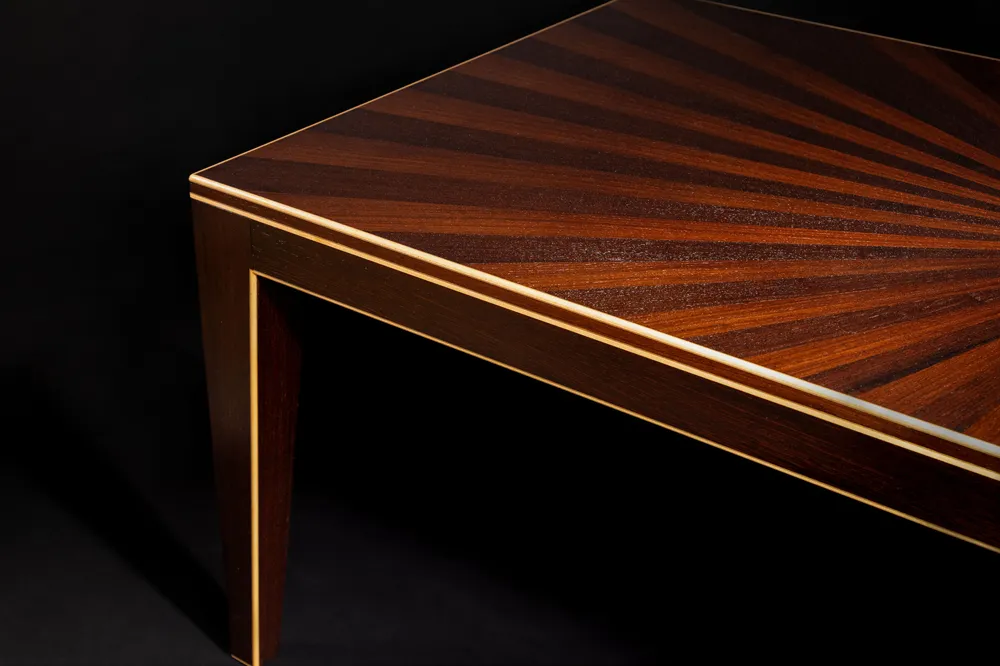 Close-up of a wooden table corner with a sunburst pattern on the tabletop and light-colored inlay trim.