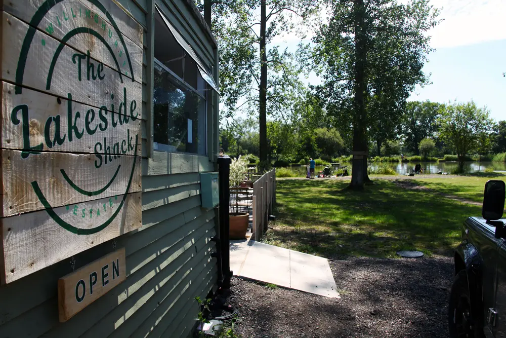 Wooden sign on a building reading 'The Lakeside Shack' and an 'OPEN' sign, with a grassy lakeside area and people in the background.