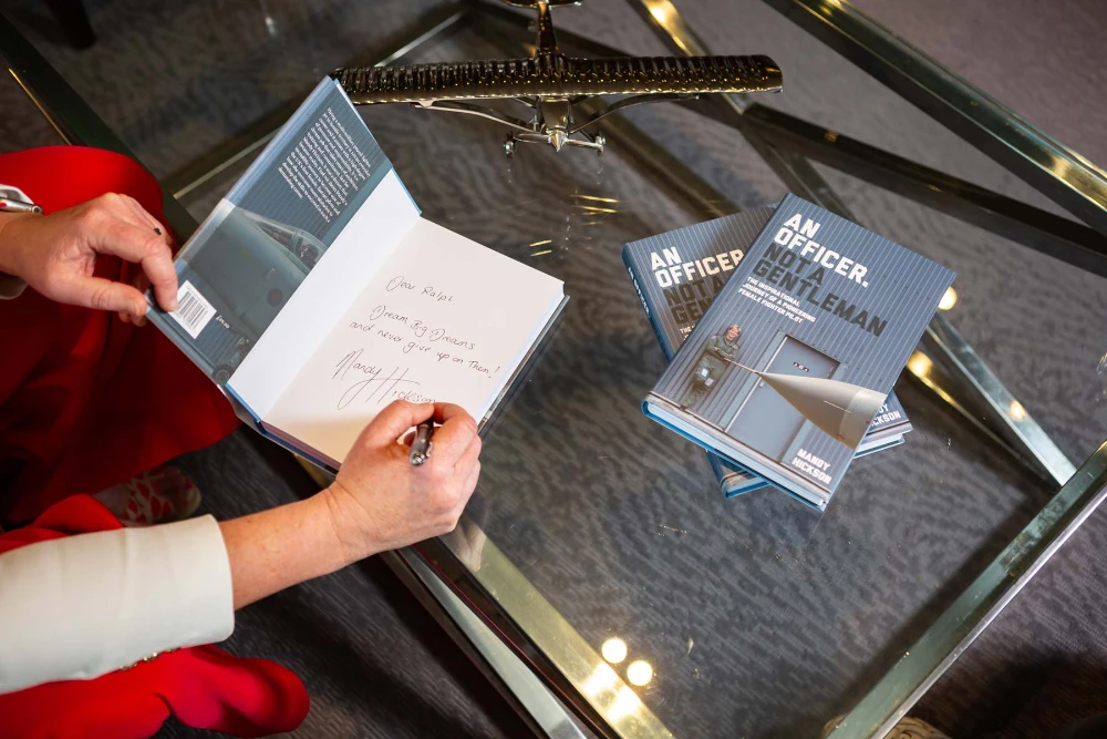 Mandy Hickson signing the inside cover of the book titled 'An Officer, Not a Gentleman' by Mandy Hickson, with two more copies on a glass table.
