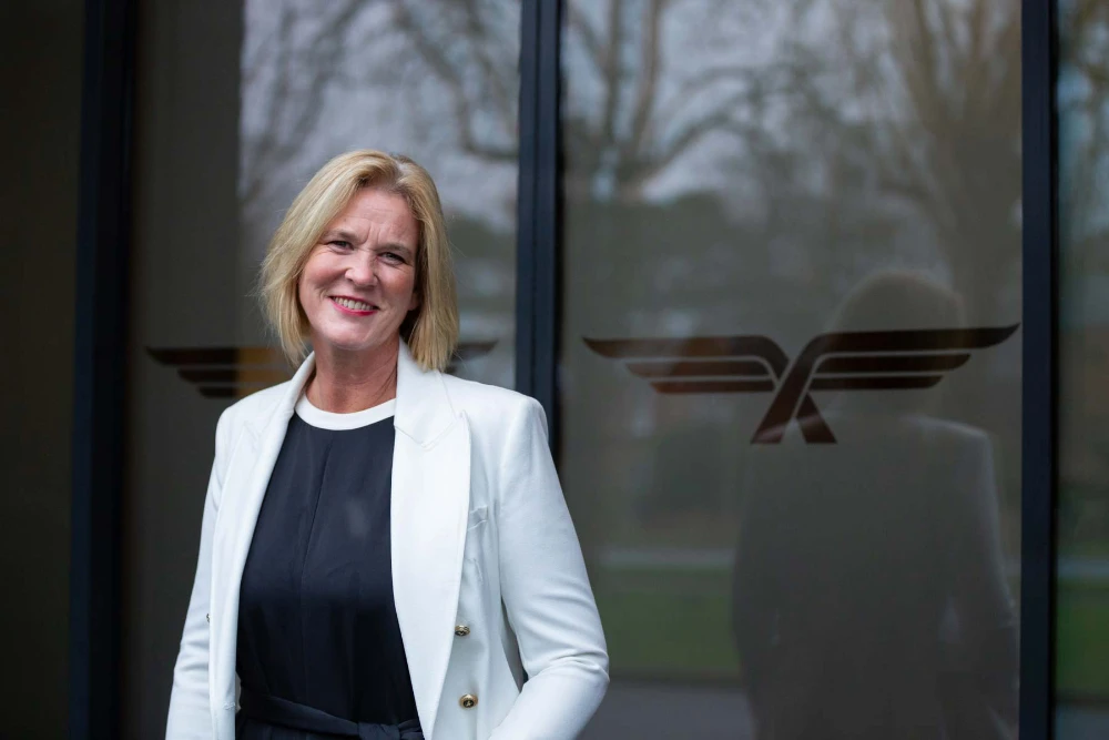 Mandy Hickson smiling wearing a white blazer stands in front of glass doors with a winged logo.