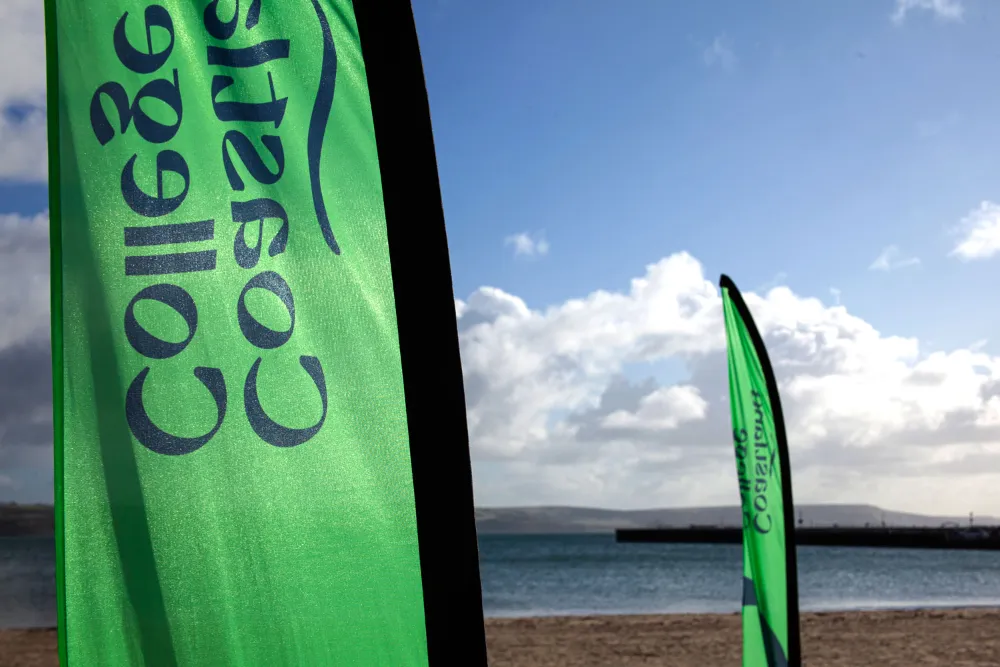 Two green Coastland College branded flags on a sandy beach.