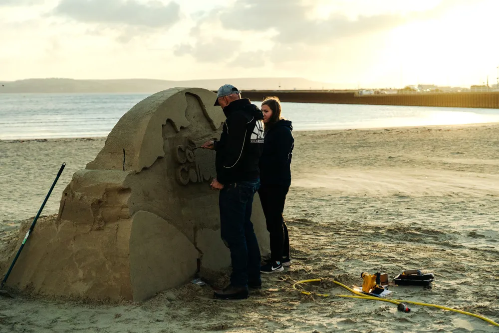 Two people sculpting a large detailed sandcastle on a beach near the ocean at sunset.