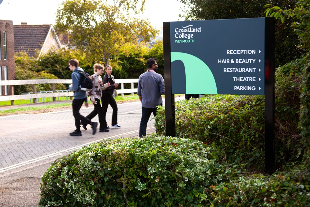 A Coastland College directional sign in blue, white and green. There are people walking around in the background.