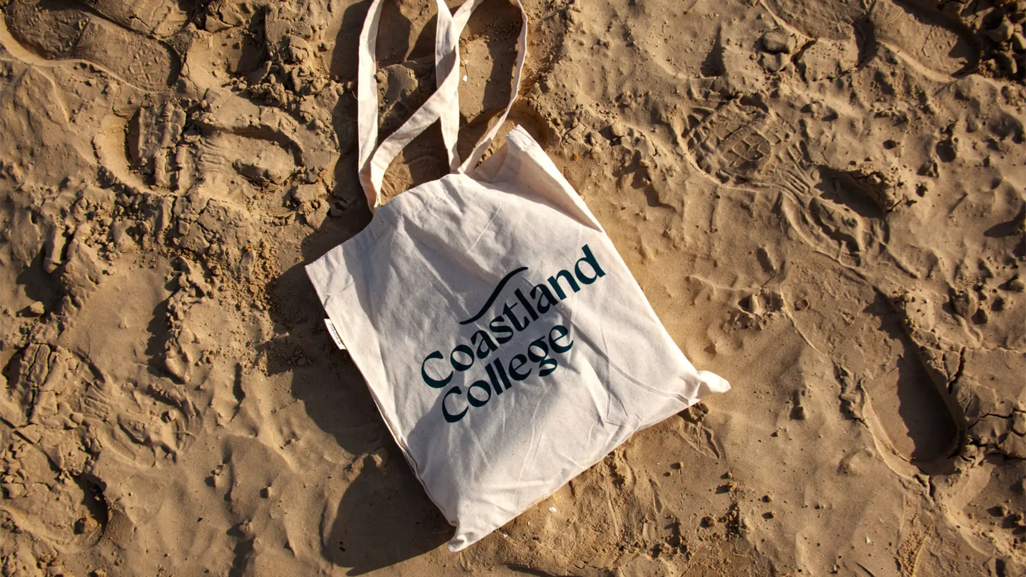 Coastland College branded tote bag laid out on a sandy beach.